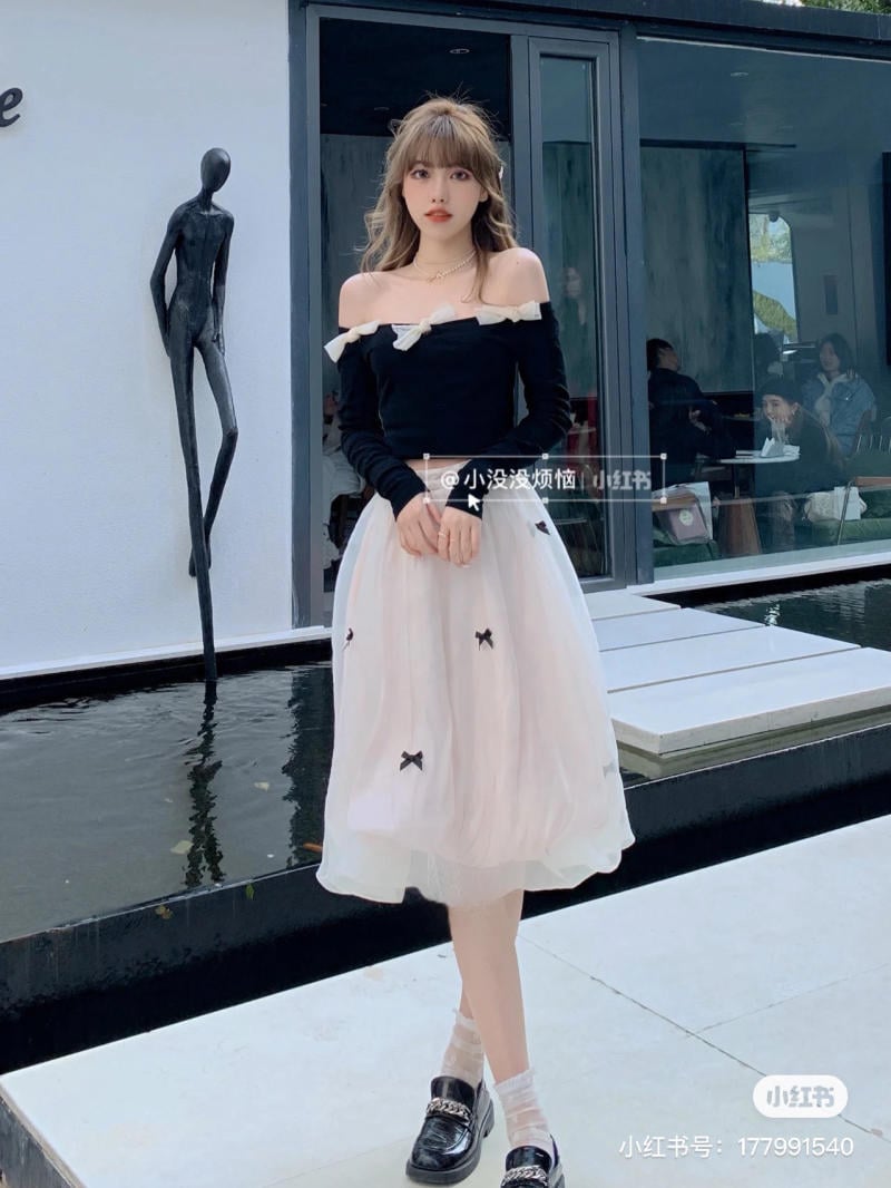 Người phụ nữ mặc váy đen trắng đứng cạnh tượng trừu tượng, phía sau là cửa kính lớn và hồ nước.