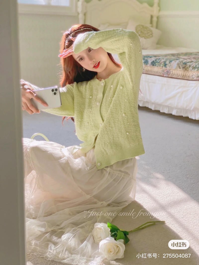 Người phụ nữ mặc áo len xanh nhạt, váy trắng, đang chụp ảnh selfie trước gương trong phòng ngủ.