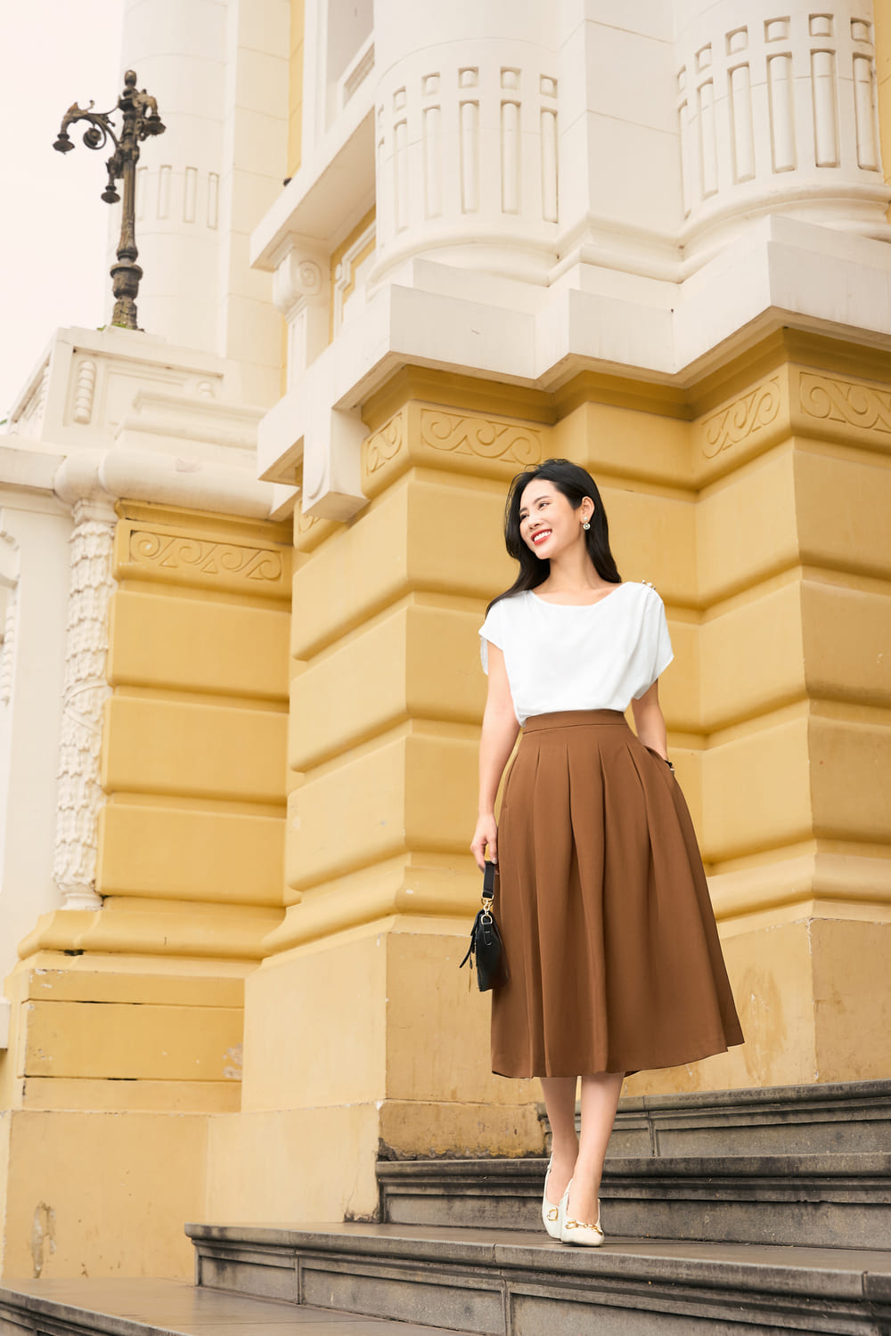 Người phụ nữ mặc váy nâu đứng trên bậc thềm trước một tòa nhà kiến trúc cổ điển màu vàng.