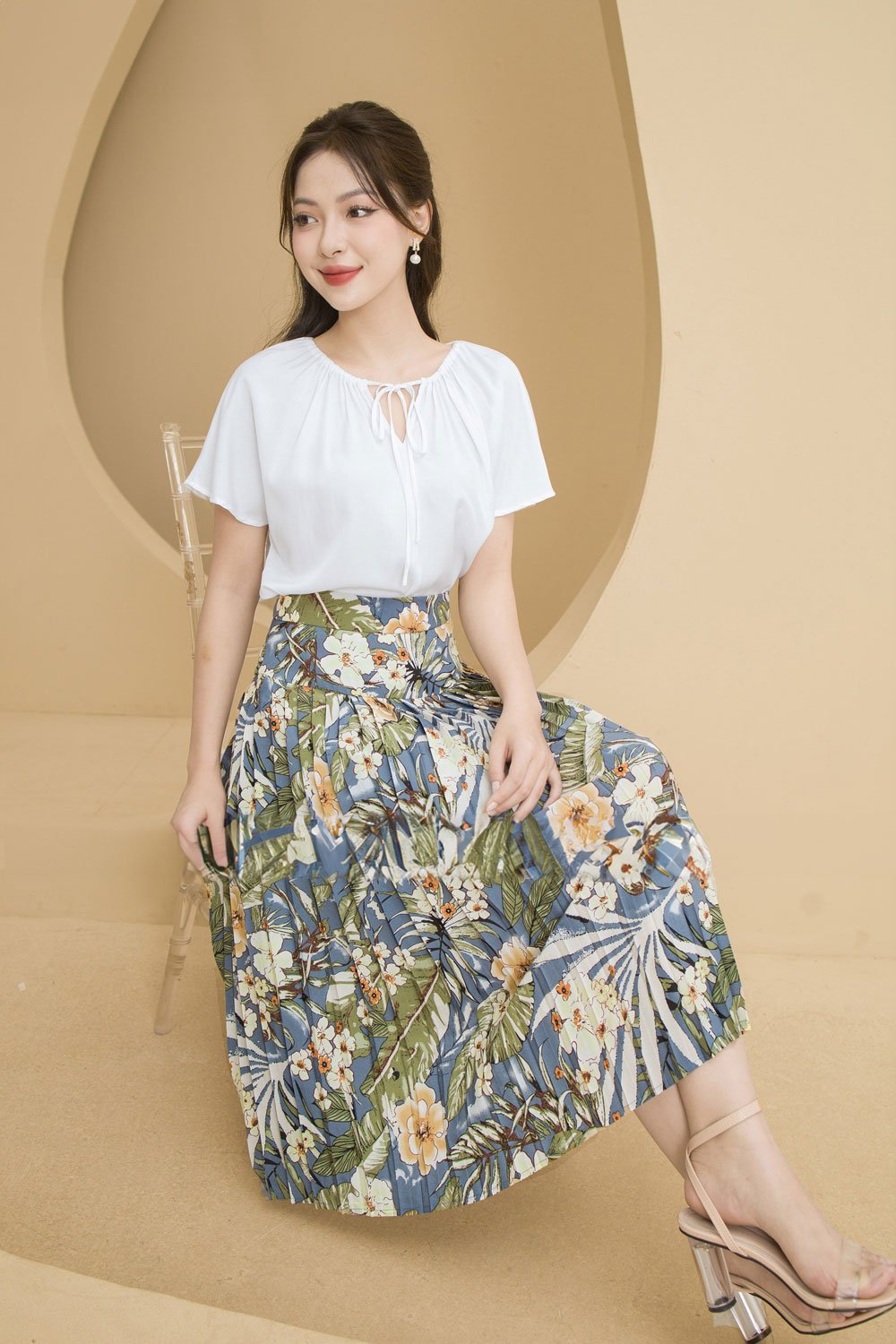 10 Cách Phối Đồ Với Chân Váy Hoa Nhí Giúp Bạn Tỏa Sáng Mùa Hè 12 Người phụ nữ mặc áo trắng và váy hoa ngồi trên ghế trong không gian nền màu be.