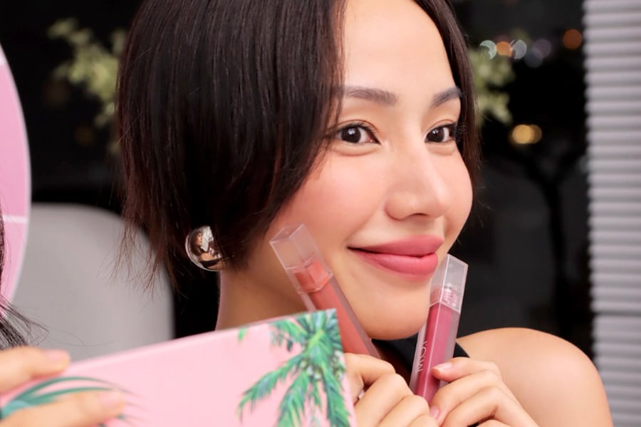 Người phụ nữ cầm hai thỏi son, mỉm cười trước ống kính, nền tối với ánh sáng lấp lánh phía sau.