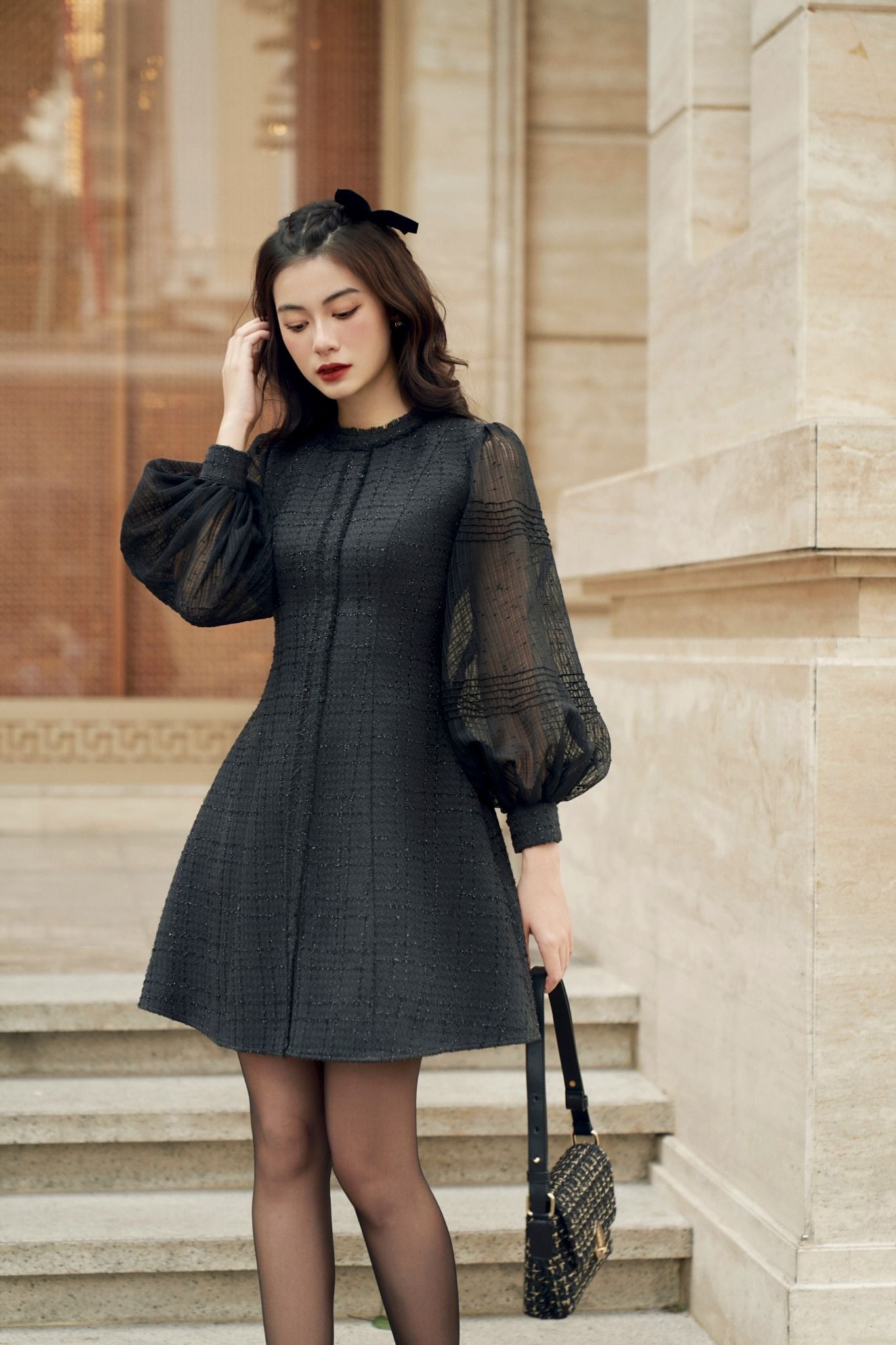 Người phụ nữ mặc váy đen đứng trên cầu thang, tay cầm túi xách, tóc dài gợn sóng, nền là tường đá.