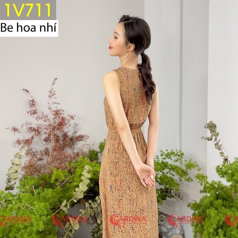 Người phụ nữ mặc váy dài hoa văn đứng cạnh cây xanh trong phòng, quay lưng về phía máy ảnh.