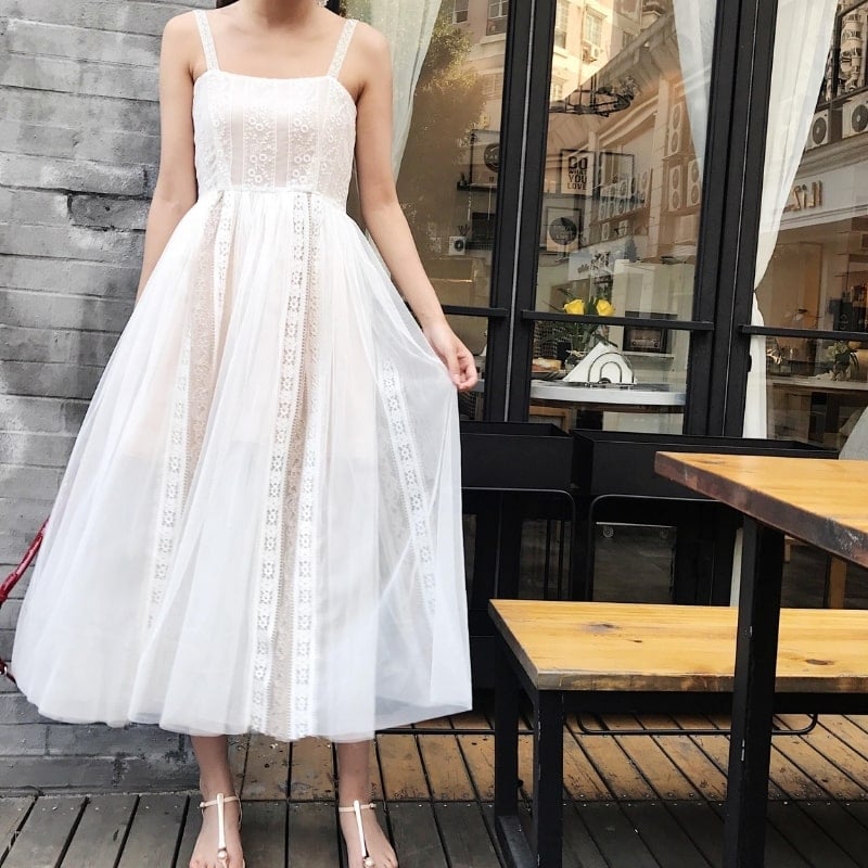 Người mặc váy trắng ren đứng trên sân gỗ cạnh bàn, nền là cửa kính và tòa nhà.