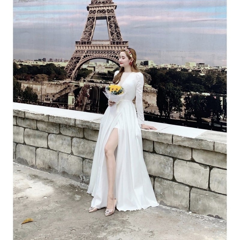 Người mặc váy trắng cầm bó hoa, đứng trước bức ảnh tháp Eiffel trên bức tường gạch.