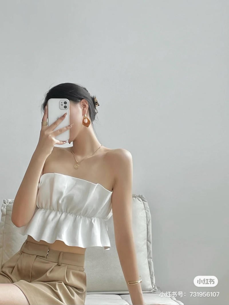 Người cầm điện thoại che mặt, mặc áo trắng và váy nâu, ngồi trên ghế sofa màu sáng.
