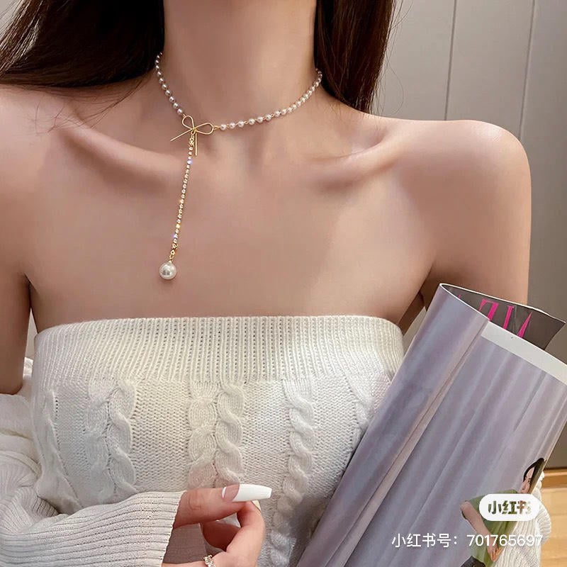 Người mặc áo len trắng và dây chuyền ngọc trai, cầm một cuốn tạp chí cuộn tròn trên tay.