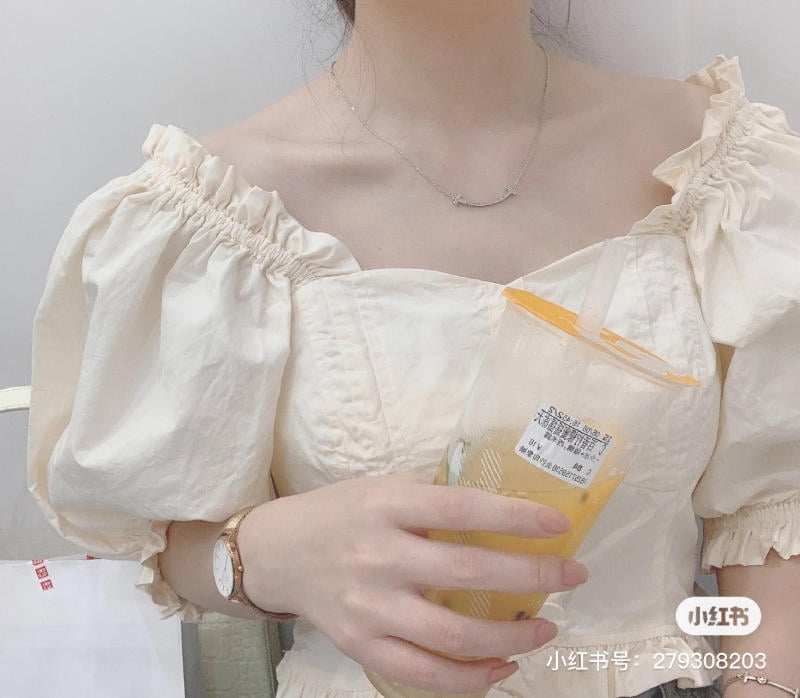 Người mặc váy trắng cầm một ly nước màu vàng, đeo đồng hồ và vòng cổ, không nhìn thấy khuôn mặt.