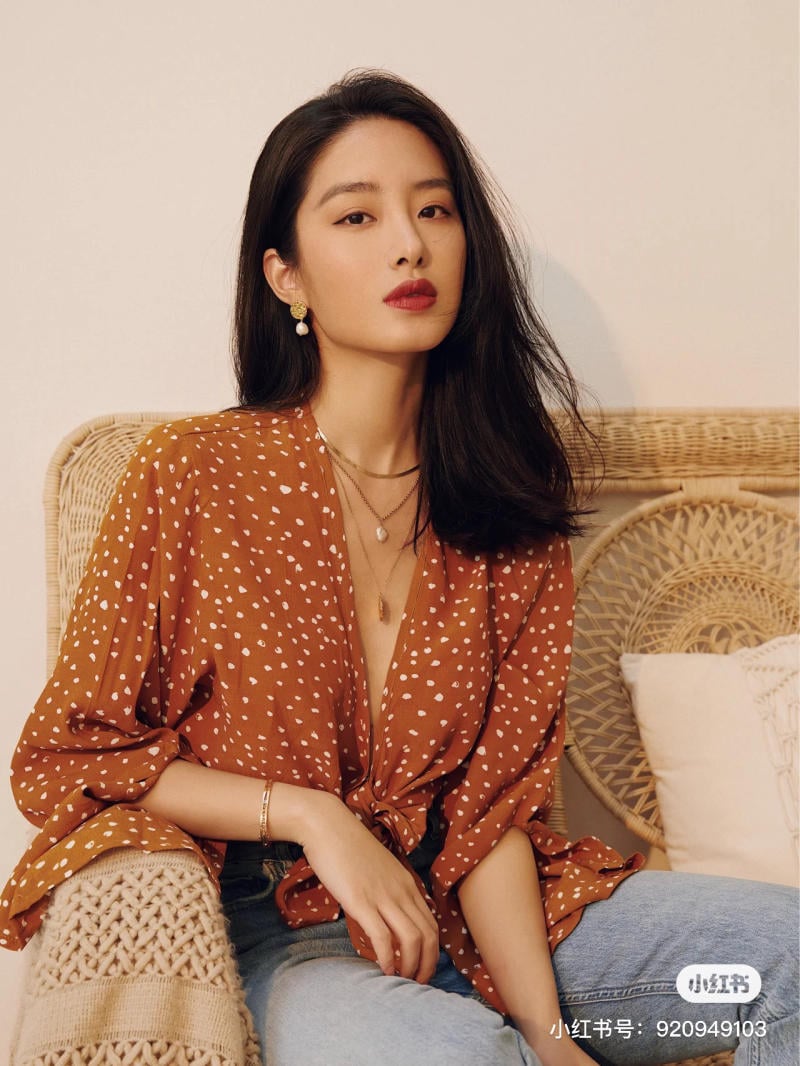 Người phụ nữ mặc áo chấm bi nâu, ngồi trên ghế mây, tóc dài xõa, đeo trang sức vàng, nhìn thẳng.