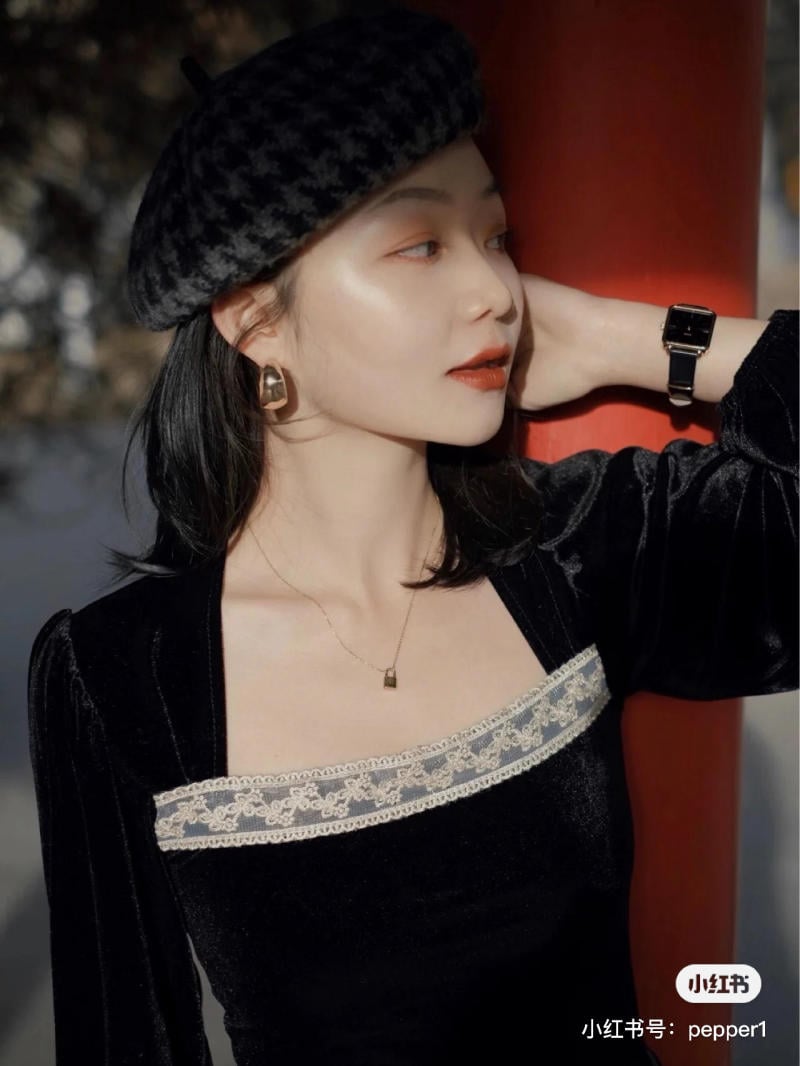 Người phụ nữ đội mũ nồi đen, mặc áo nhung đen viền ren, đeo đồng hồ và bông tai lớn, nhìn sang bên.