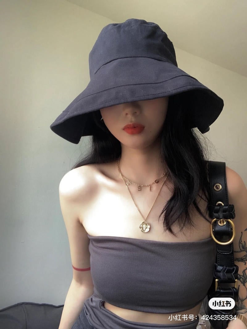Người đội mũ rộng vành, mặc áo quây màu xám, đeo dây chuyền vàng và túi xách đen bên vai.