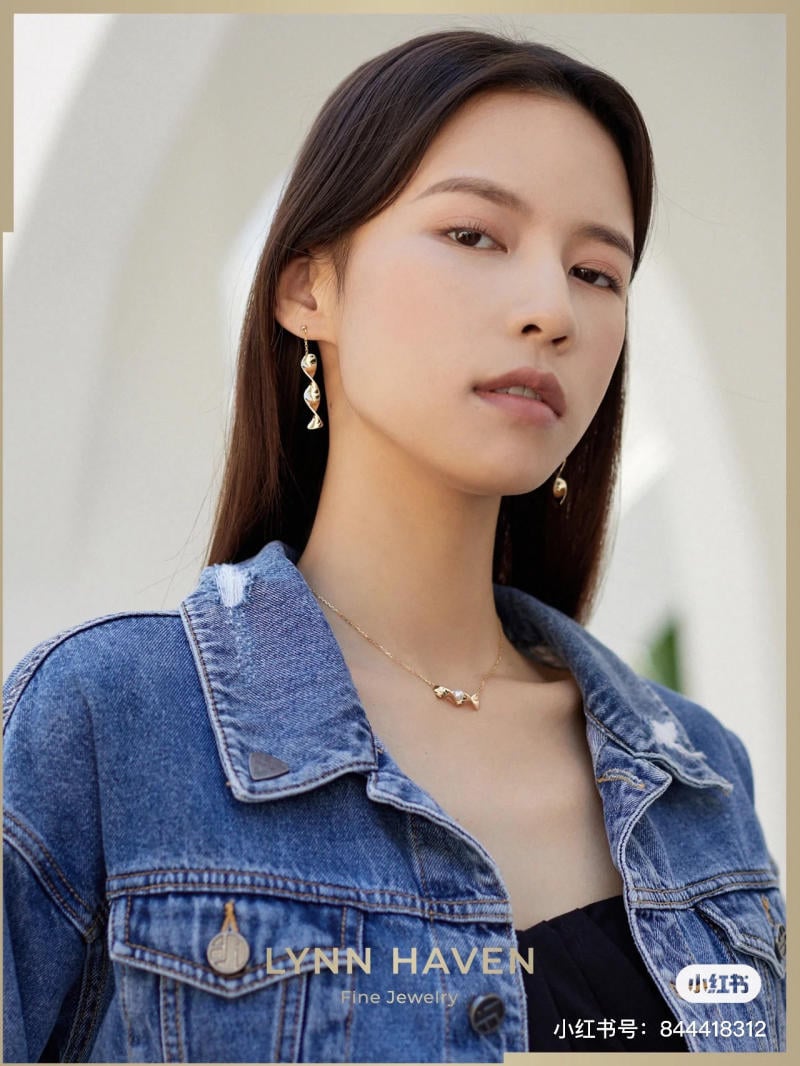 Người mẫu mặc áo khoác denim, đeo trang sức vàng, đứng trước nền sáng.