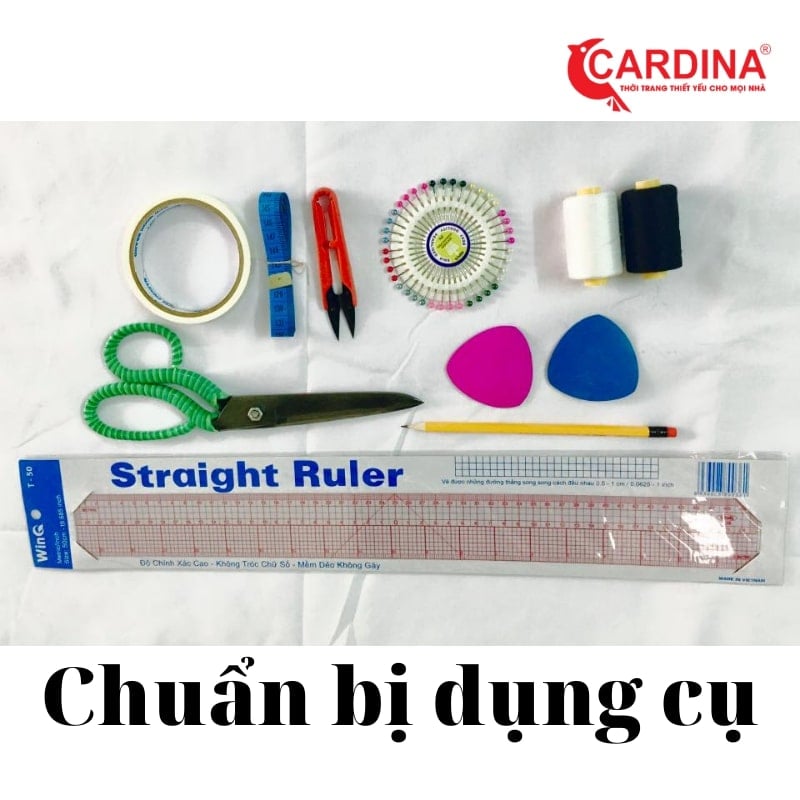 Các dụng cụ may vá bao gồm kéo, thước, băng dính, phấn may, kim ghim, chỉ và bút chì trên nền vải trắng.