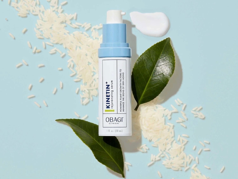 d7a230bd-serum-cho-da-hon-hop-thien-dau-14 Chai serum trên nền xanh, xung quanh có lá trà xanh và hạt gạo.