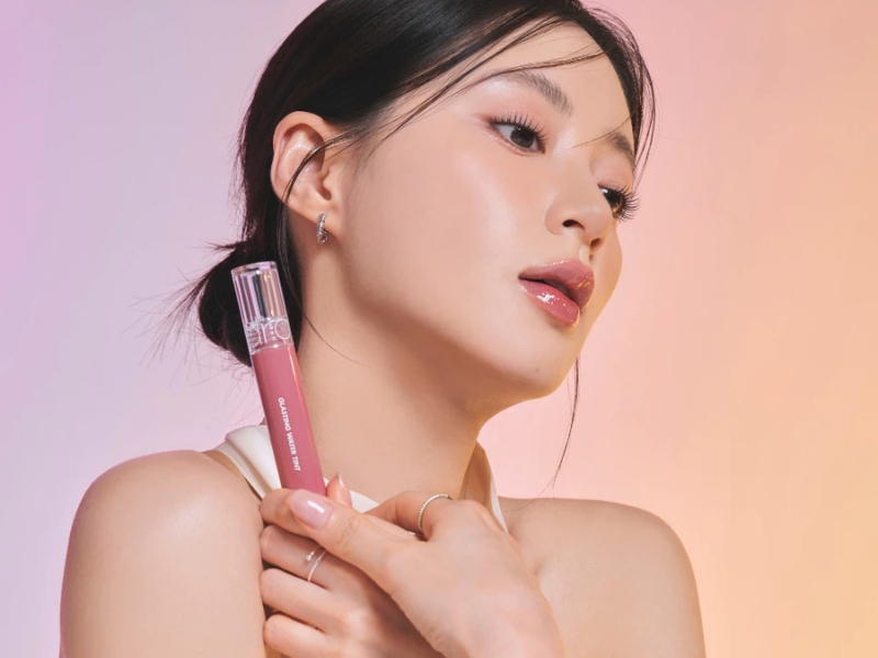 Người phụ nữ cầm lọ son môi màu hồng, nền màu sắc nhẹ nhàng, phong cách trang điểm tự nhiên.