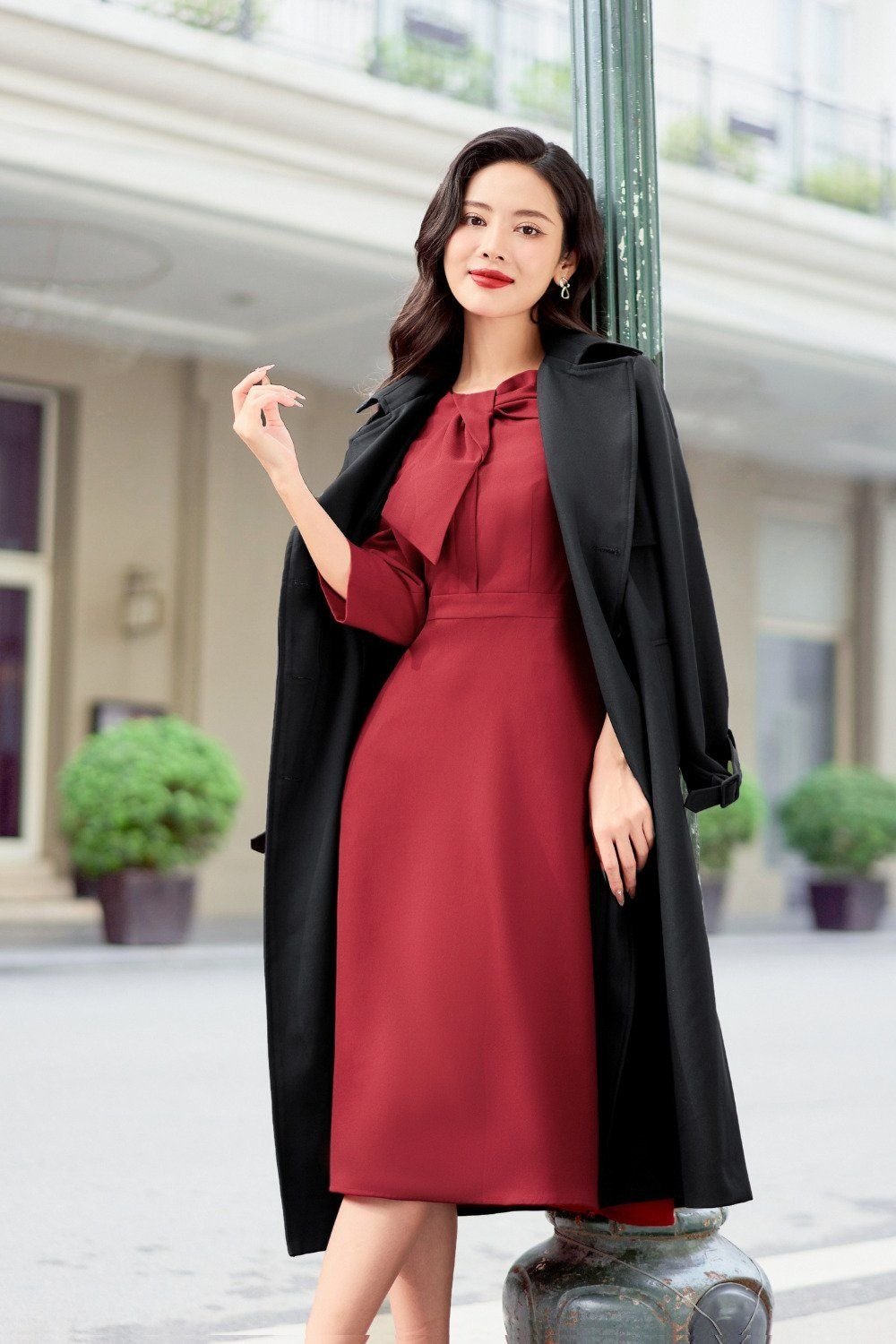 Người phụ nữ mặc váy đỏ và áo khoác đen đứng cạnh cột đèn trên phố, phông nền có tòa nhà và cây xanh.