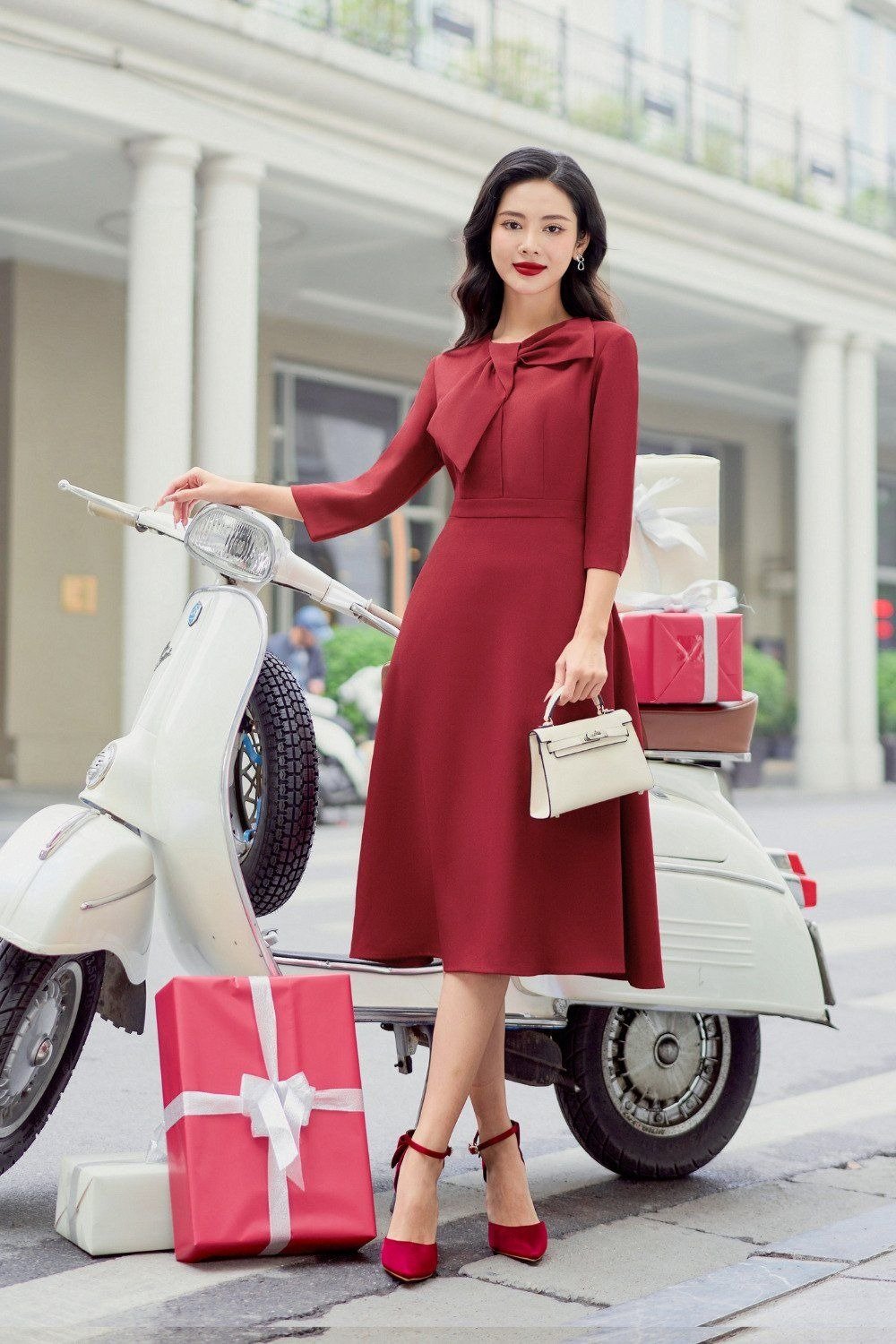 Người phụ nữ trong trang phục đỏ đứng cạnh xe máy trắng, xung quanh có nhiều hộp quà màu đỏ và trắng.