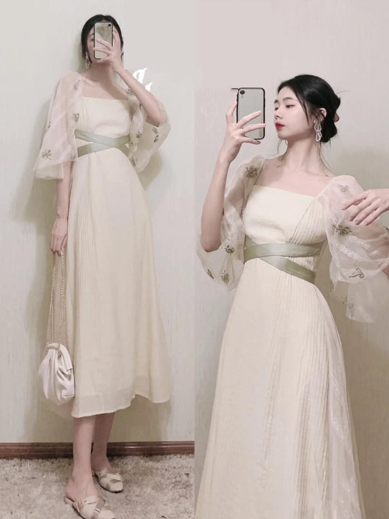 Người mặc váy trắng chụp ảnh trước gương, tay cầm điện thoại, mặc áo ngoài xuyên thấu thêu hoa.