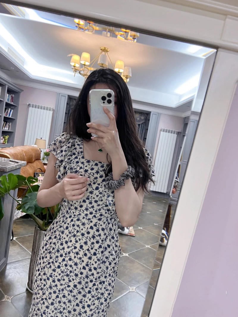 Người chụp ảnh tự sướng trong gương, mặc váy hoa, cầm điện thoại, trong phòng khách với đèn chùm.