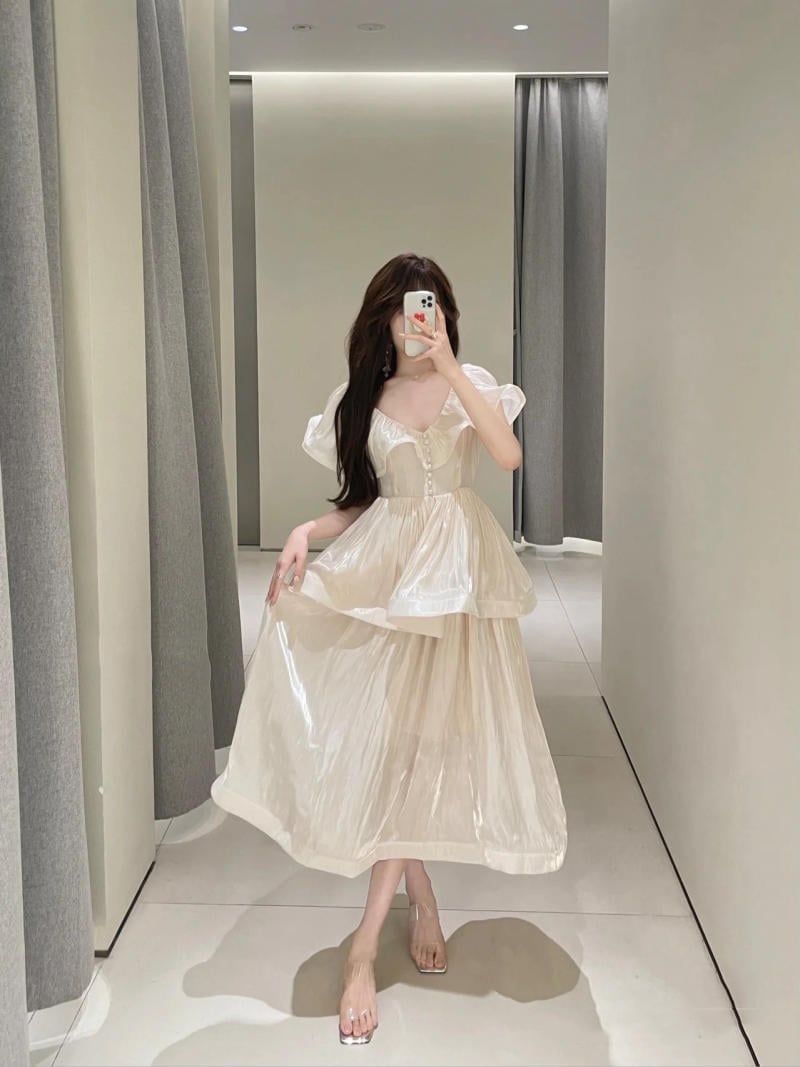 Người chụp ảnh tự sướng trong gương, mặc váy trắng dài, đứng trong phòng có rèm và ánh sáng dịu.