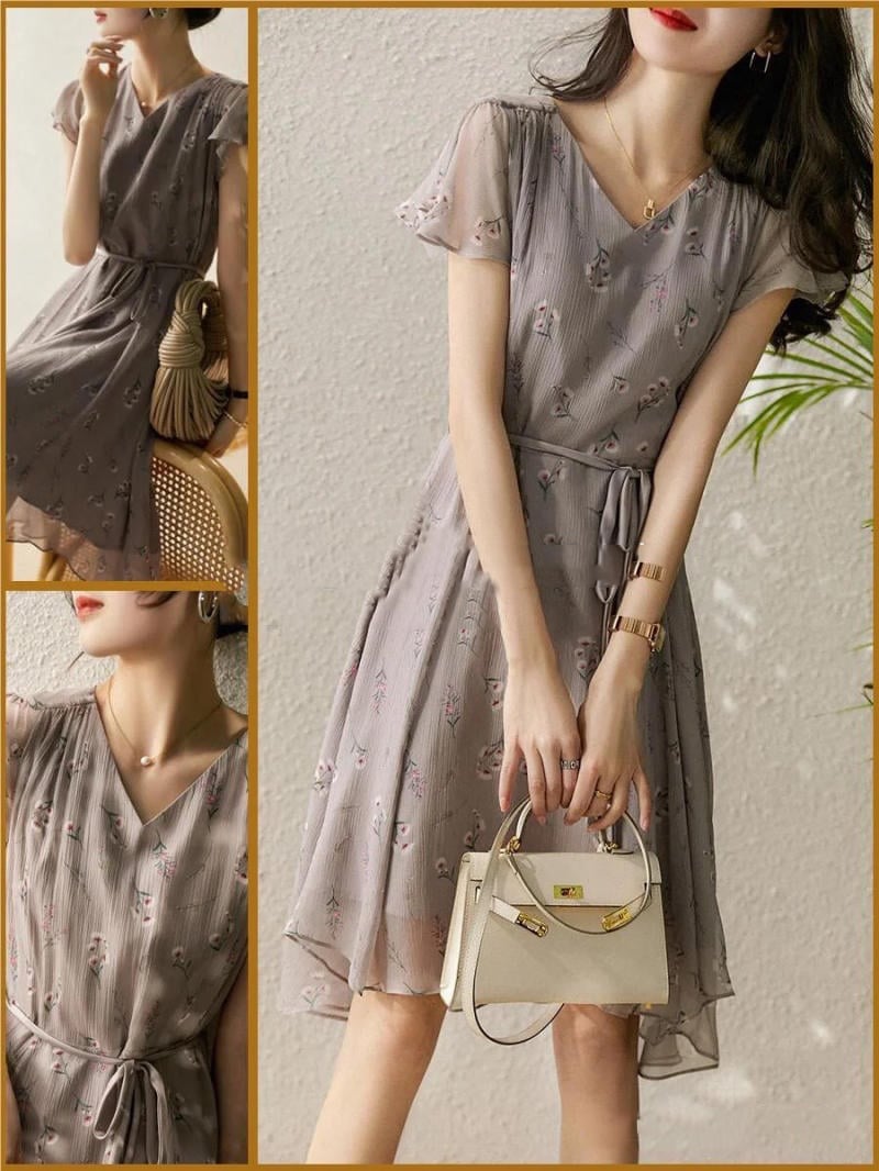 Người mẫu mặc váy hoa màu xám, cầm túi xách nhỏ màu trắng, tạo dáng trước nền tường sáng.
