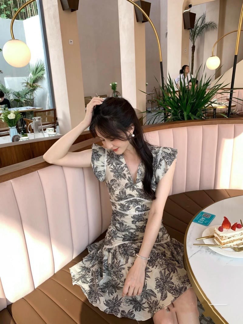 Người phụ nữ mặc váy hoa ngồi trong quán cà phê, trên bàn có bánh ngọt và trang trí cây xanh.