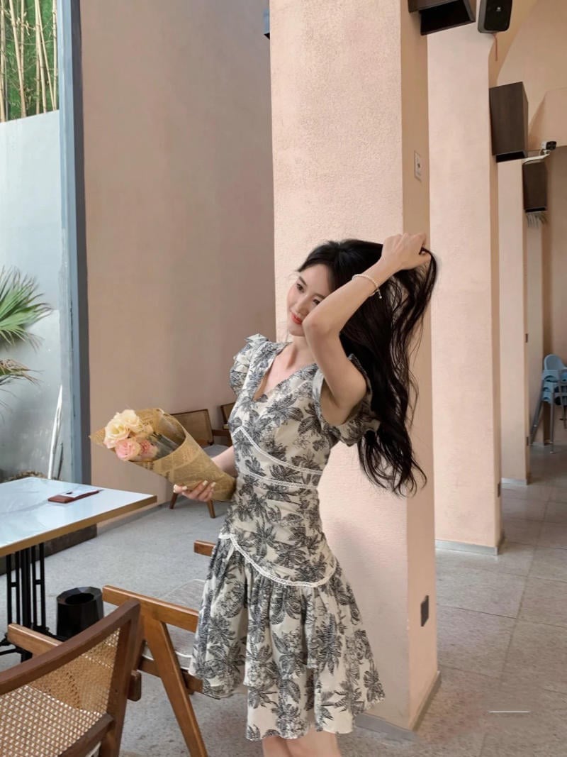 Người cầm bó hoa đứng trong không gian nội thất, mặc váy họa tiết hoa, tay vuốt tóc.