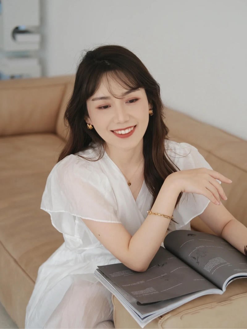 Người phụ nữ mặc váy trắng ngồi trên ghế sofa, cầm cuốn tạp chí mở và mỉm cười.