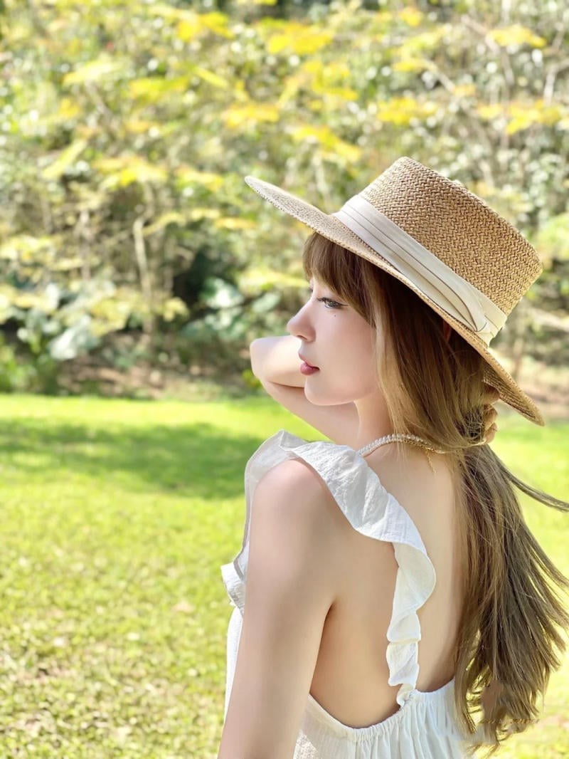 Người phụ nữ đội mũ rơm, mặc váy trắng, đứng ngoài trời trong khu vườn xanh tươi đầy nắng.