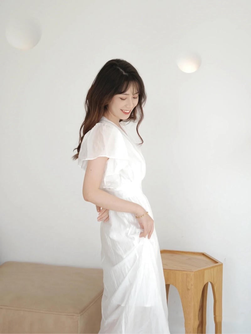 Người phụ nữ mặc váy trắng, nghiêng đầu cười, đứng cạnh bàn gỗ trong phòng sáng.