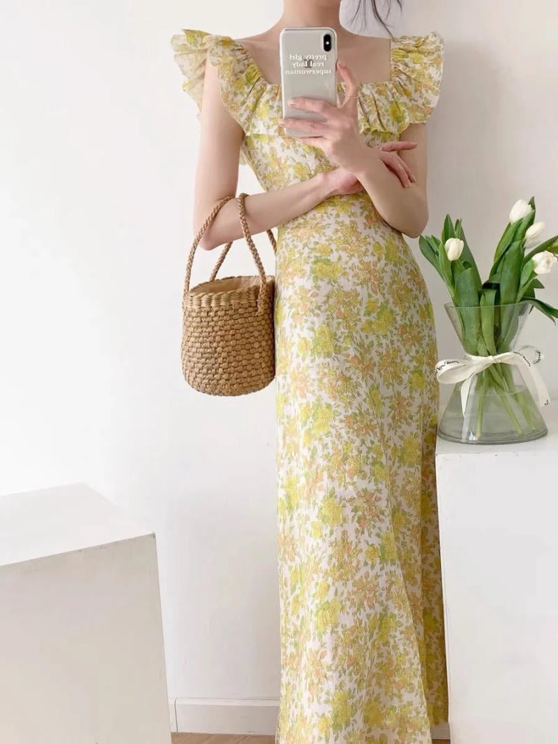 Người mặc váy hoa dài, cầm điện thoại và túi cói, đứng cạnh bình hoa tulip trắng trong phòng sáng sủa.