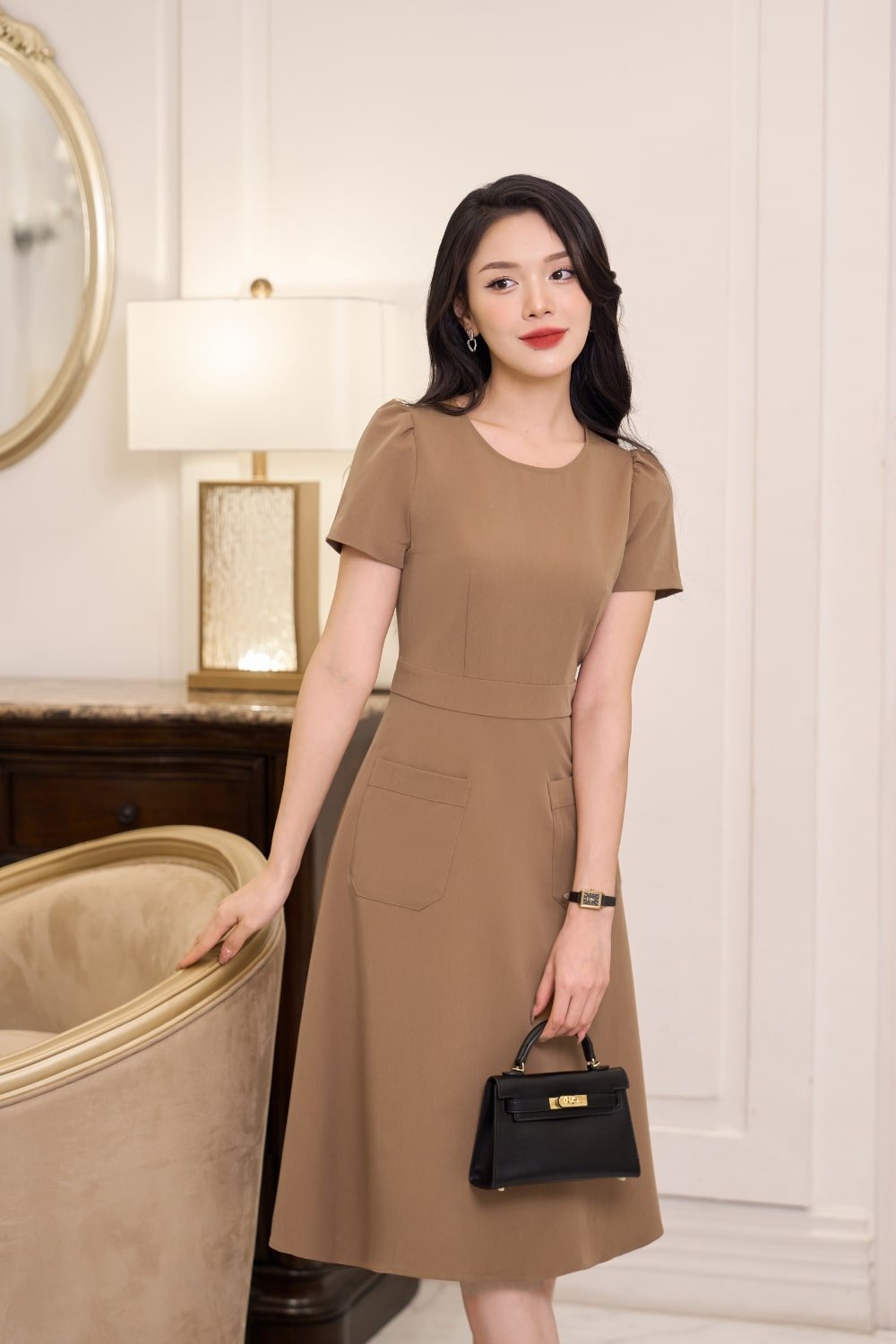 dam-chu-a__2__9550b4ce4dbf46aea50a4654f6f00799 Người phụ nữ mặc váy nâu, cầm túi xách đen, đứng cạnh ghế và đèn trong phòng có tông màu sáng.