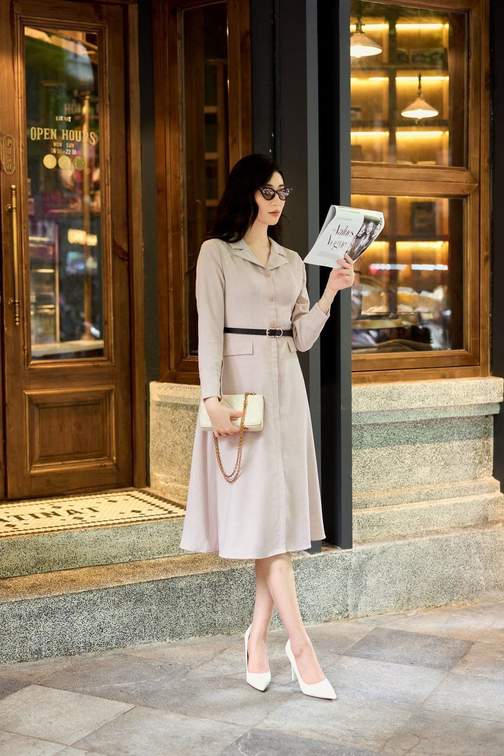 Người phụ nữ mặc váy dài, đeo kính và cầm tạp chí, đứng trước cửa quán cà phê với ánh sáng ấm áp.