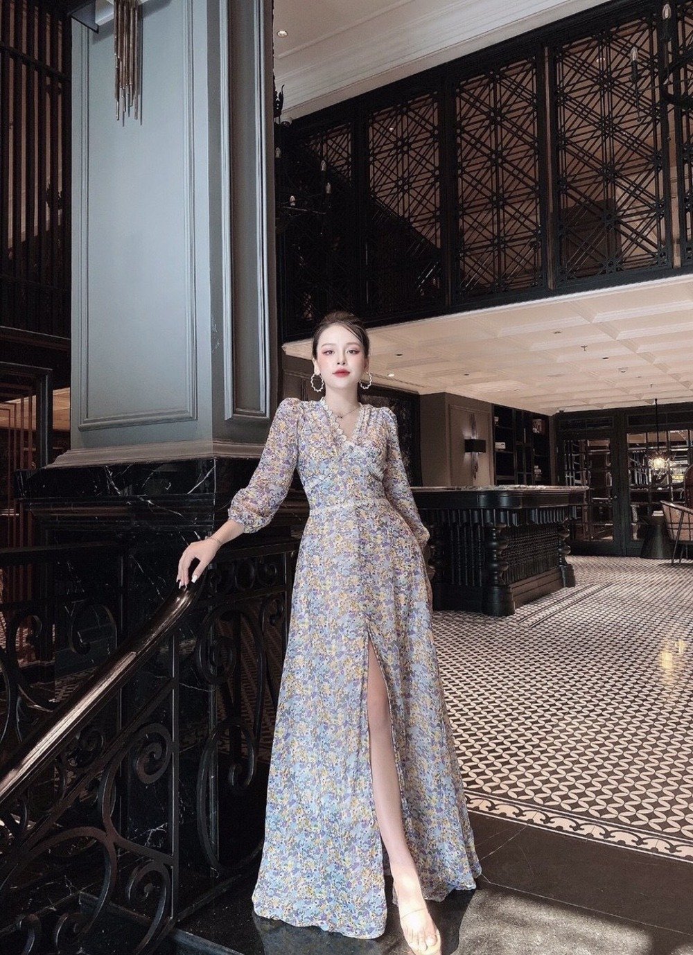 Người phụ nữ mặc váy hoa dài đứng tựa vào lan can trong không gian nội thất sang trọng.