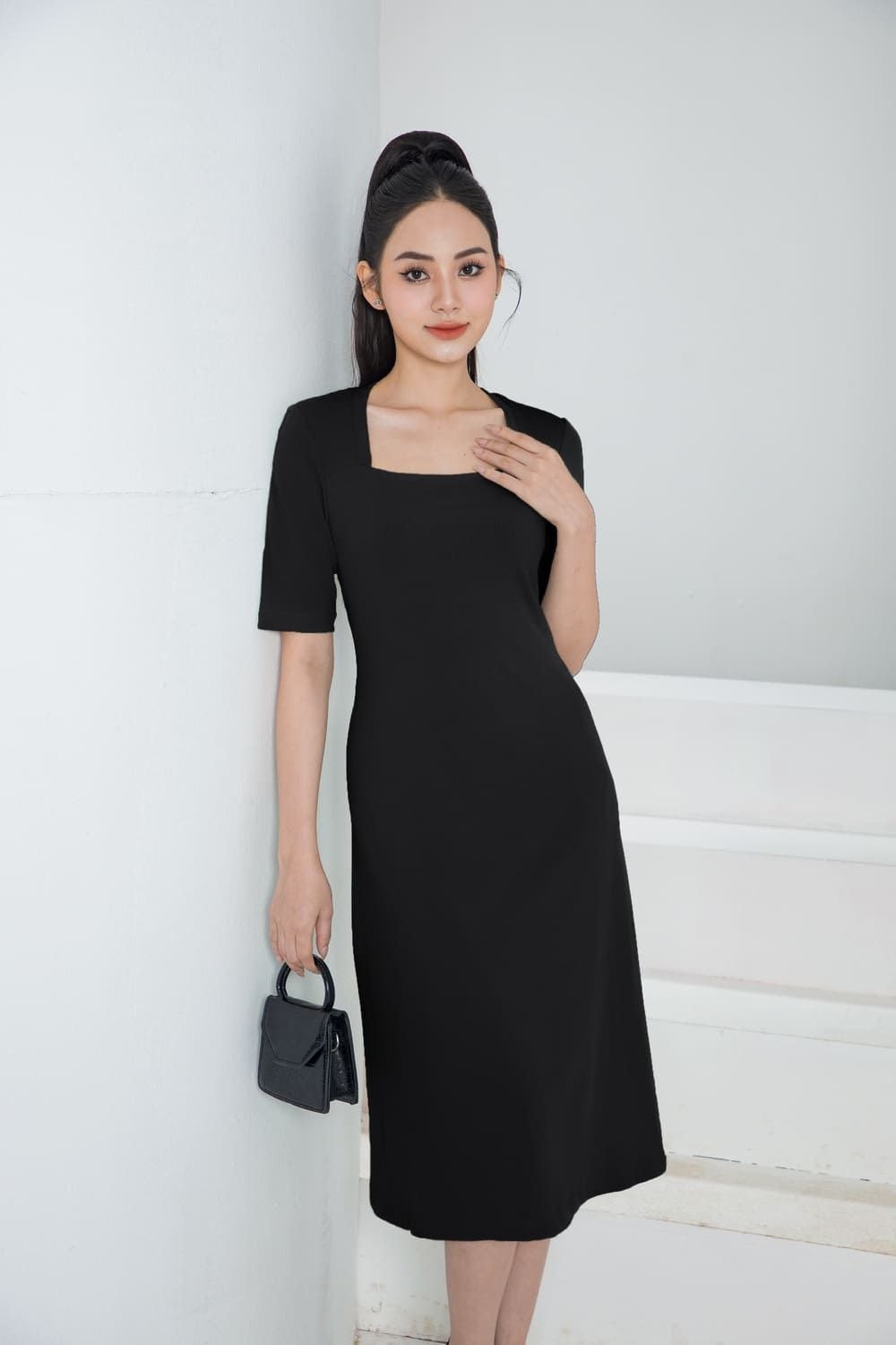 Người phụ nữ mặc váy đen dài, đứng dựa vào tường trắng, tay cầm túi xách, tóc buộc cao.