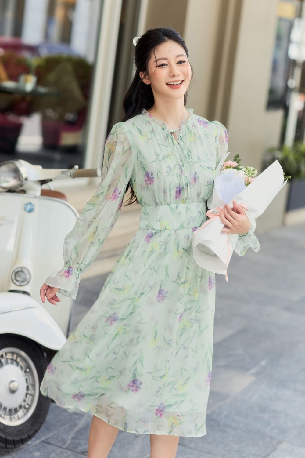 Người phụ nữ mặc váy hoa, cầm bó hoa và cười tươi trên phố, bên cạnh là một chiếc xe máy màu trắng.