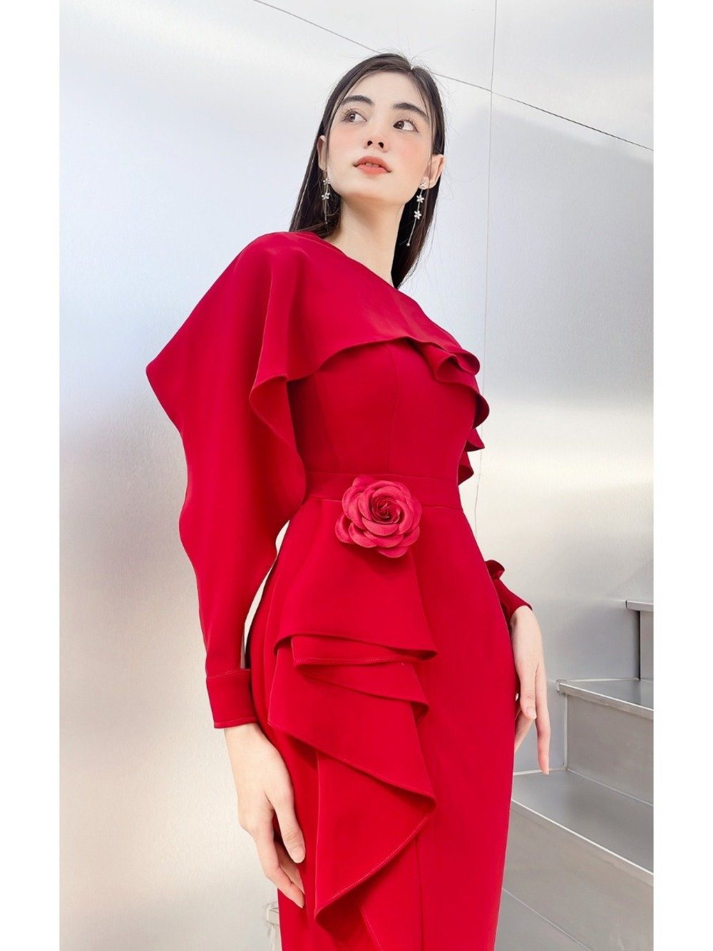 Người mẫu mặc váy đỏ rực rỡ với hoa hồng trang trí, đứng trước nền tường kim loại sáng bóng.