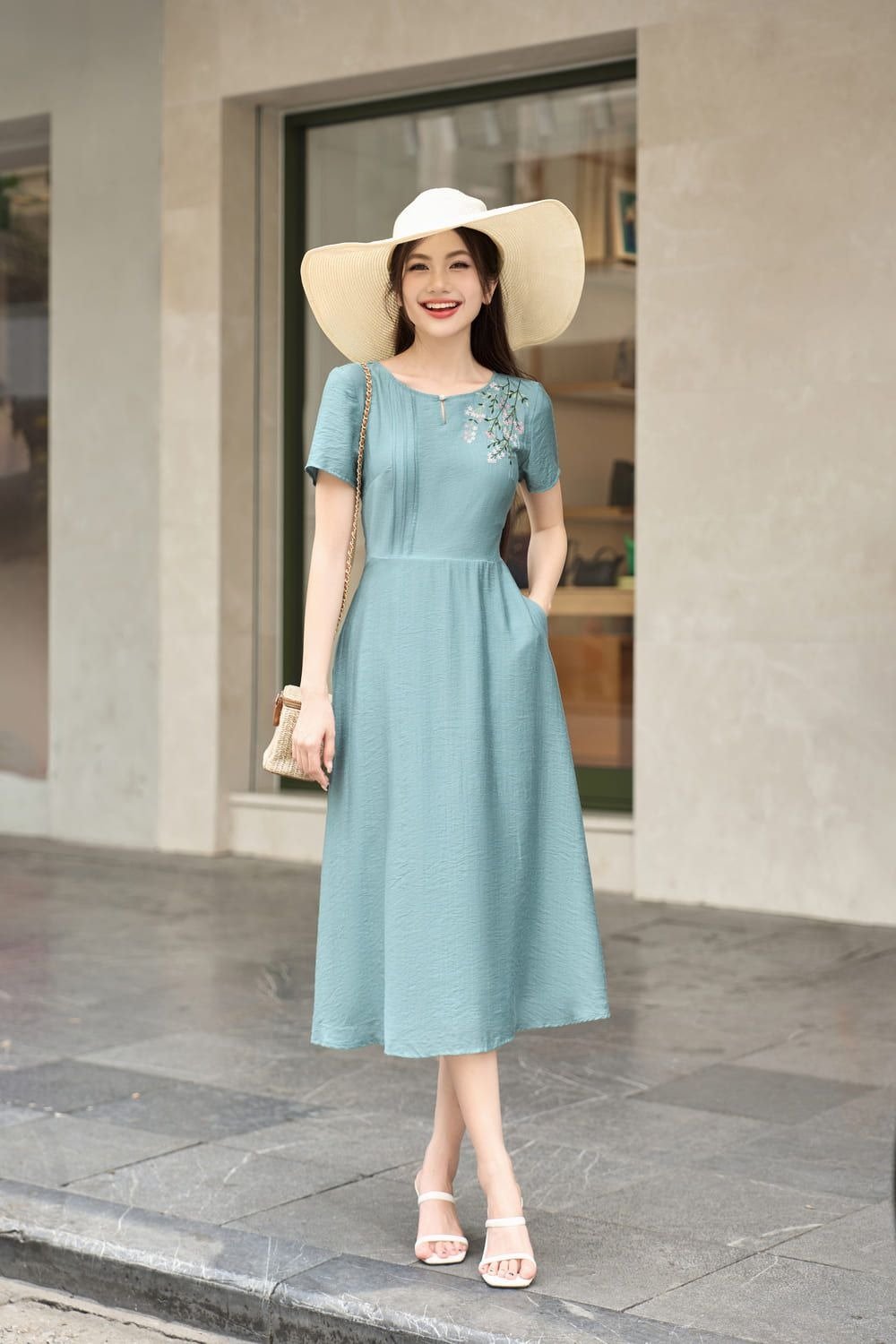 Người phụ nữ mặc váy xanh và đội mũ rộng vành đứng trước cửa hàng, cười tươi.