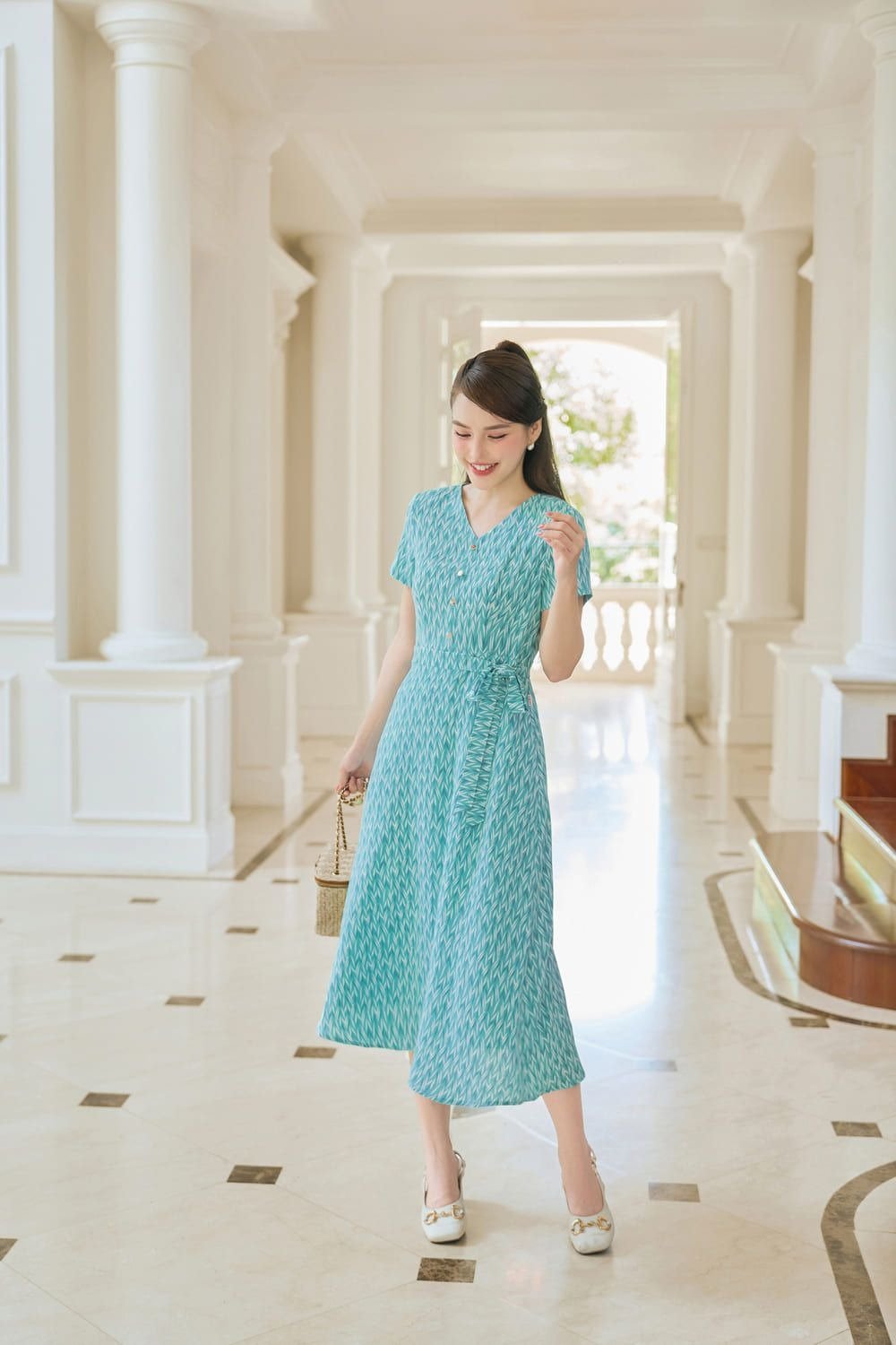 Người phụ nữ mặc váy xanh, đứng trong hành lang sáng sủa với sàn lát đá hoa.