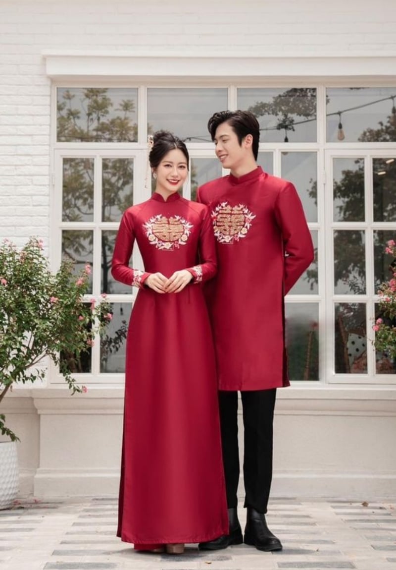 Hai người mặc áo dài đỏ truyền thống đứng cạnh nhau, phía sau là cửa sổ lớn và cây xanh.