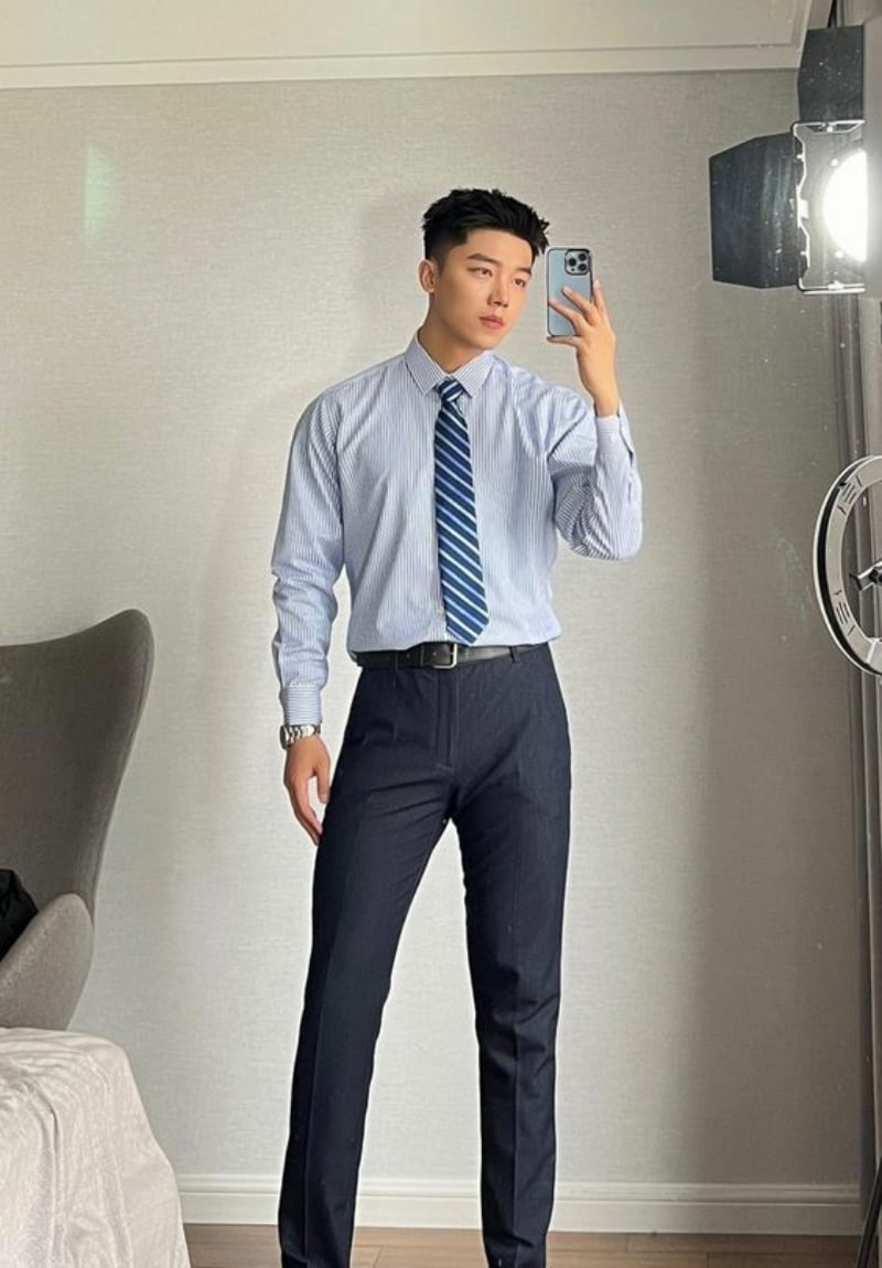Người đàn ông mặc áo sơ mi và cà vạt đứng trước gương, chụp ảnh tự sướng bằng điện thoại trong phòng.