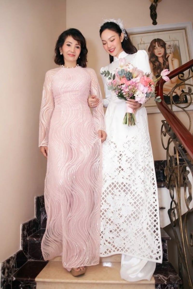 Hai người phụ nữ mặc áo dài, một người cầm hoa, đang đi xuống cầu thang trong không gian nội thất trang nhã.