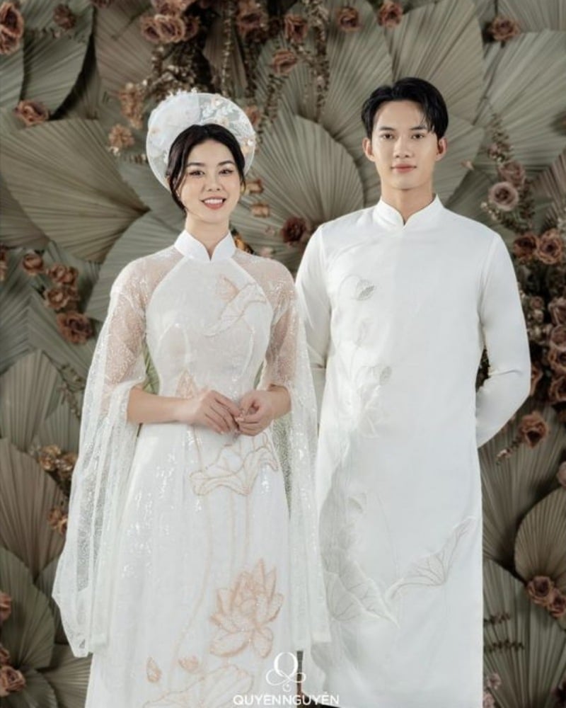 Hai người mặc áo dài trắng truyền thống, đứng trước nền trang trí hoa lá.