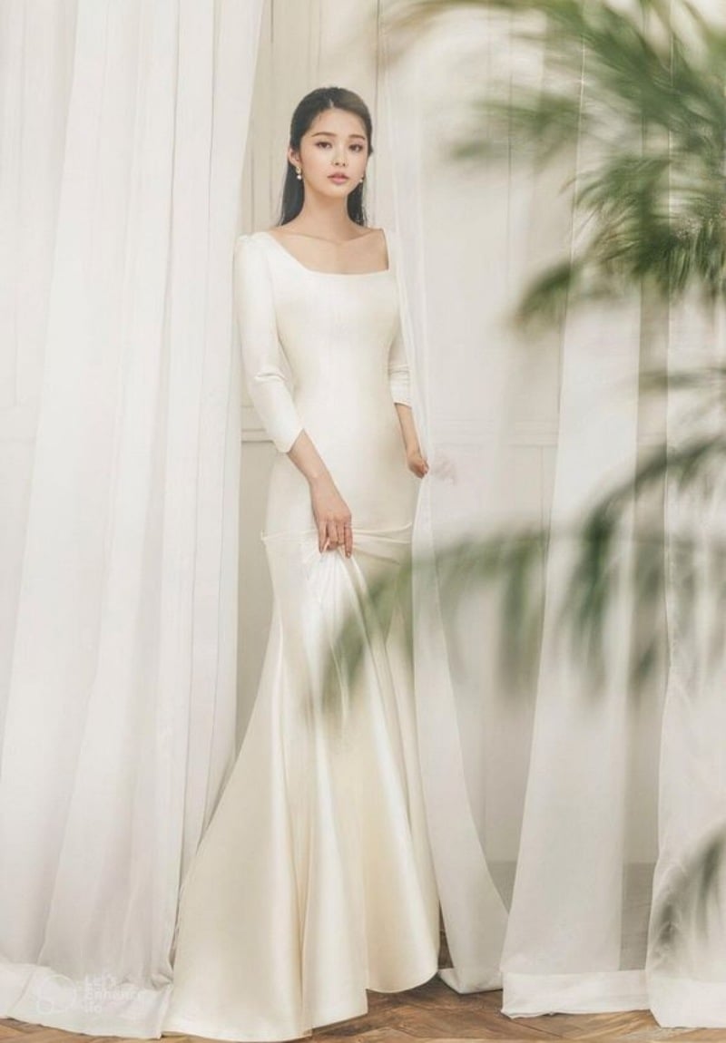 Người phụ nữ trong bộ váy trắng dài đứng cạnh rèm cửa, nền trắng, cây xanh mờ phía trước.