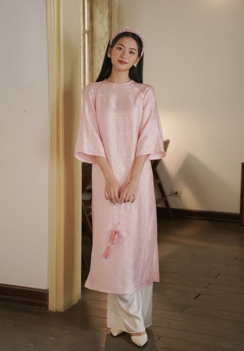 Người phụ nữ mặc áo dài hồng đứng trong phòng, tay cầm phụ kiện, ánh sáng nhẹ nhàng từ cửa sổ phía sau.