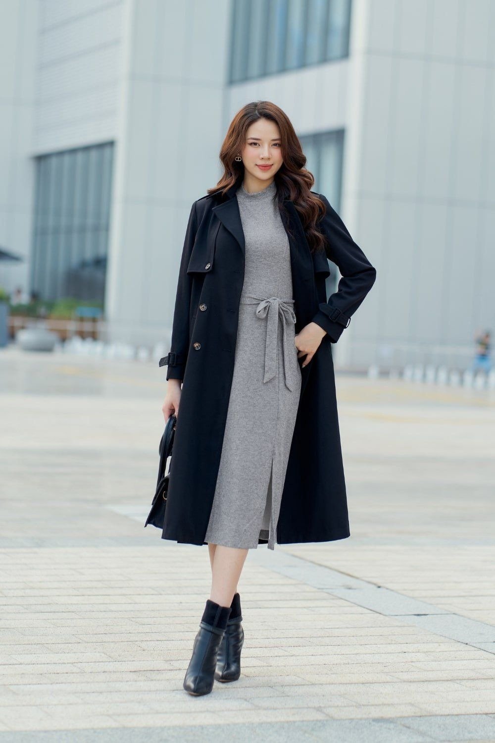 Người phụ nữ mặc váy xám và áo khoác đen, đứng trên vỉa hè trước tòa nhà hiện đại.