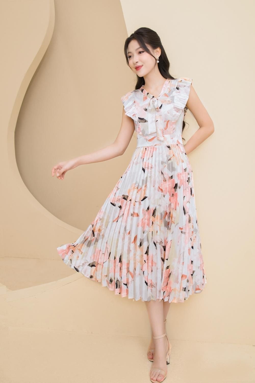 Người phụ nữ mặc váy hoa xếp li màu pastel đứng trước phông nền màu be, tạo dáng thanh lịch.