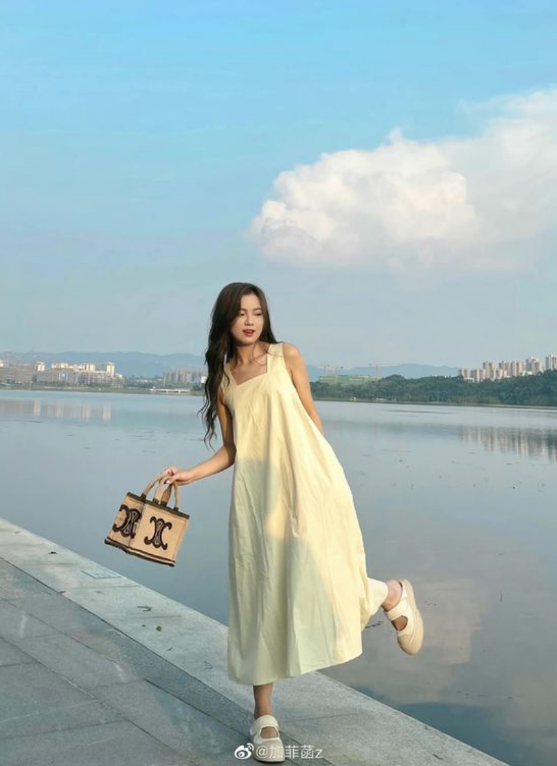 Người phụ nữ mặc váy vàng đứng cạnh hồ, xách túi, với cảnh quan thành phố và bầu trời xanh phía sau.
