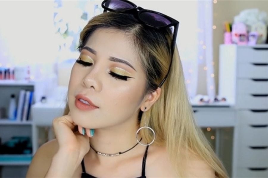 Người phụ nữ ngồi trong phòng, đeo kính râm trên đầu, trang điểm mắt nổi bật và đeo hoa tai tròn.