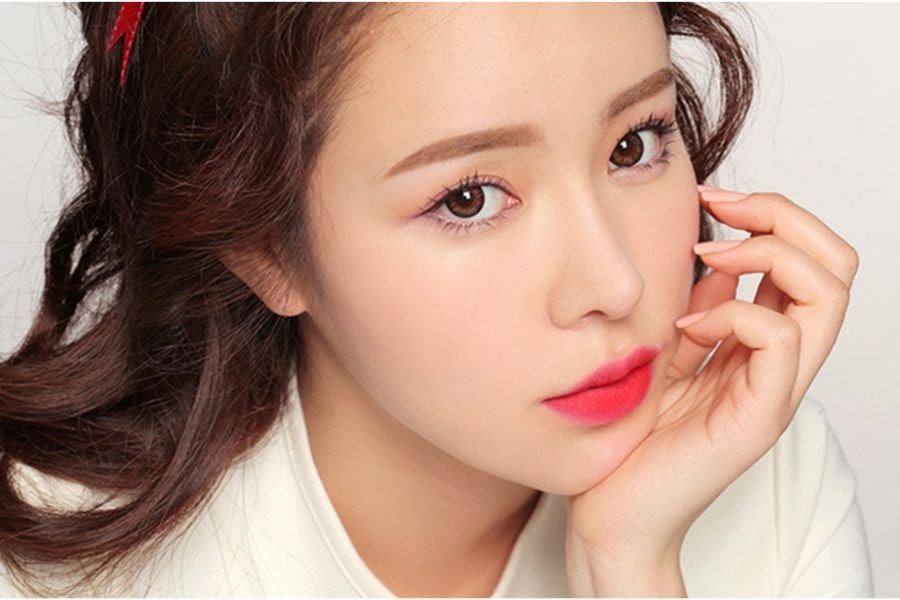Người phụ nữ nhìn thẳng vào máy ảnh, tay chạm nhẹ vào mặt, môi đỏ, tóc nâu xoăn nhẹ, nền trắng.